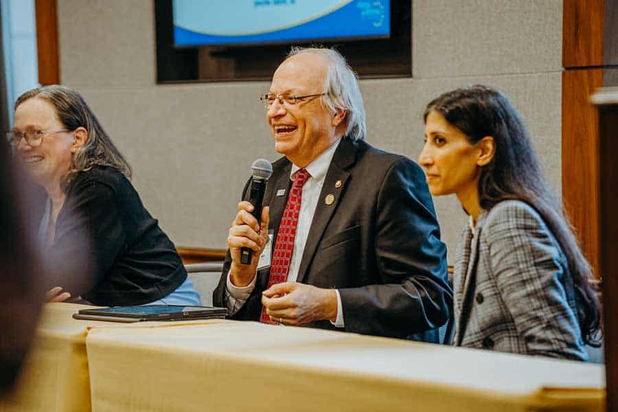 Three people sit at a table during a panel discussion. The man in the center, smiling and wearing a suit and red tie, holds a microphone. Two women sit on either side of him, listening attentively.