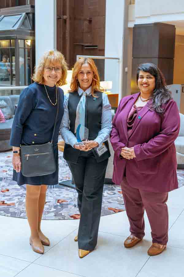 Three women stand together indoors, posing and smiling for a photo. The woman in the center holds a glass award. They are dressed in business attire and standing in a lobby area with modern decor.