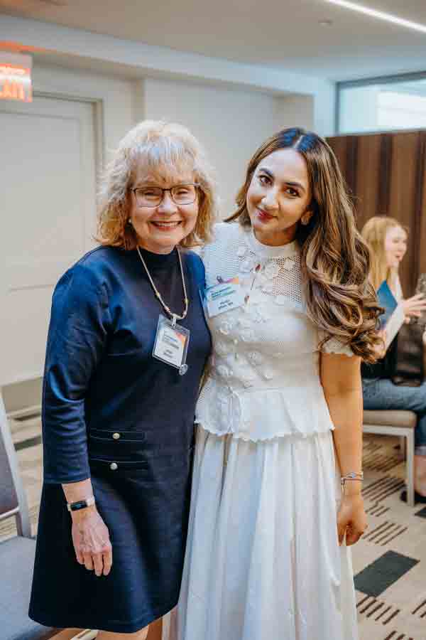 Two women smiling and standing together at an indoor event; one wears a navy dress and glasses, the other wears a white dress. Both have name tags and appear to be attending a professional or social gathering.
