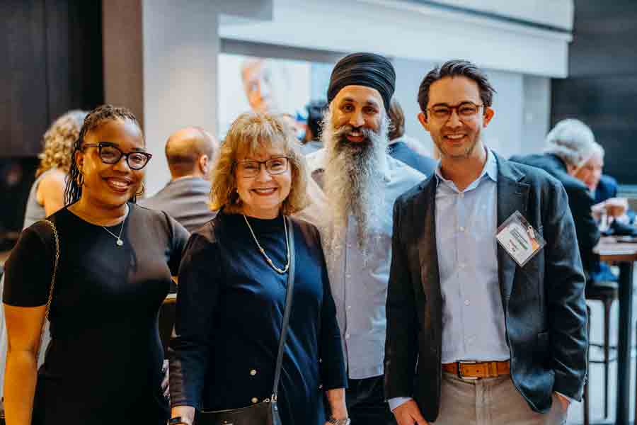 Four people stand together indoors, smiling at the camera at what appears to be a professional or social event. Others can be seen seated in the blurred background.