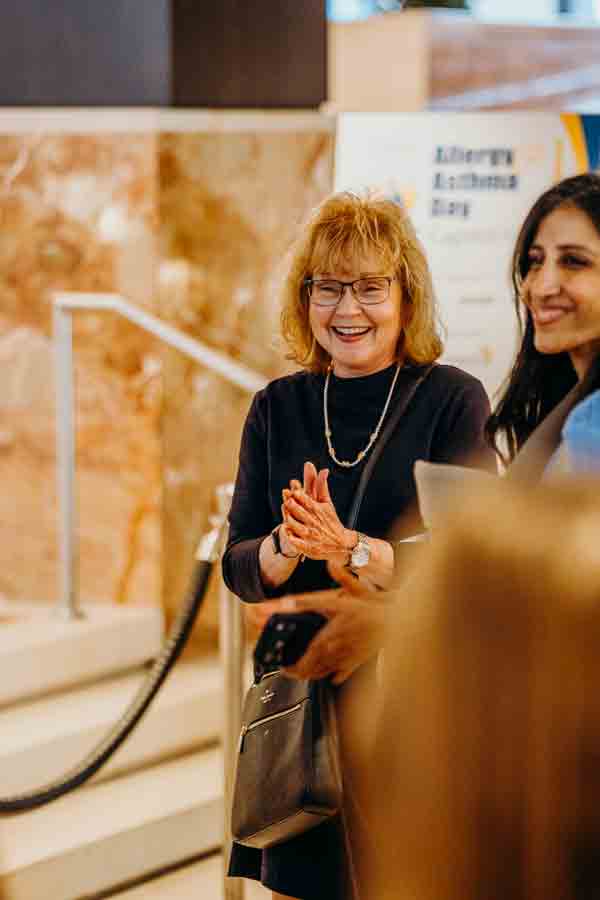 Two women stand indoors, smiling and talking. One woman with glasses and reddish hair claps her hands, while the other woman with long dark hair stands beside her. A marble wall and stairs are visible in the background.