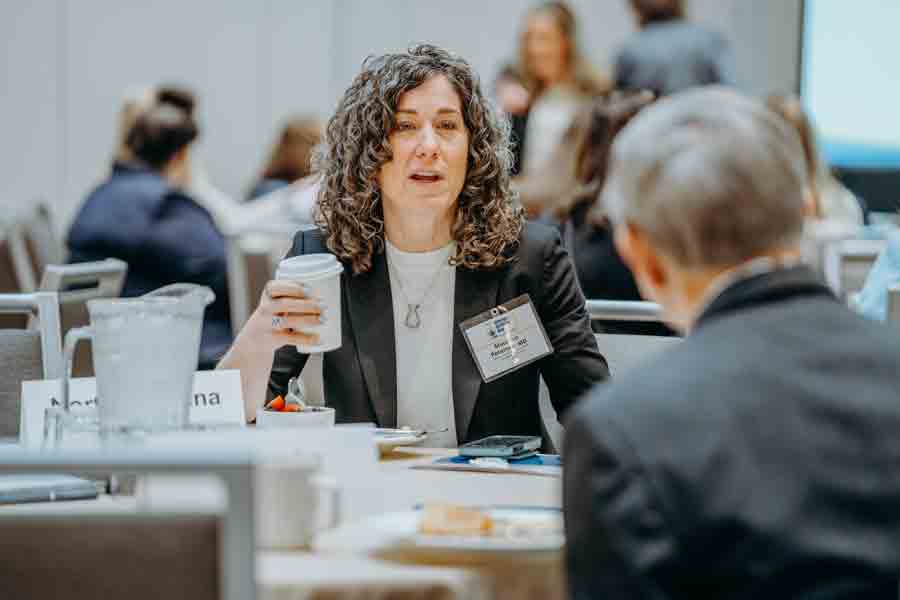 A woman with curly hair in a black blazer holds a coffee cup and speaks to a man at a conference table, with other people blurred in the background.