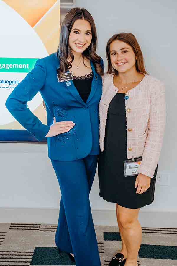 Two women stand side by side smiling at the camera. One wears a blue suit with a name badge, while the other wears a light pink jacket over a black dress. They appear to be at a professional event.