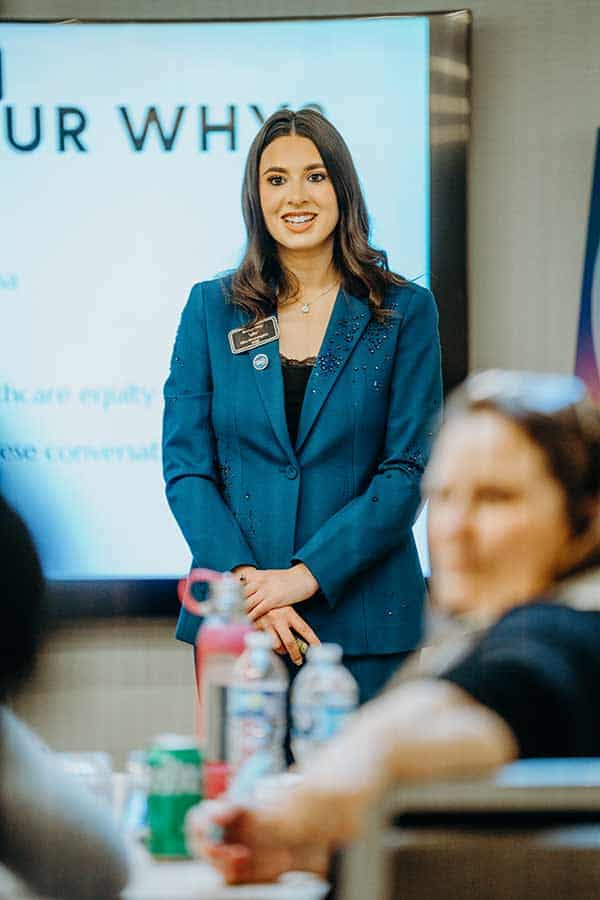 A woman in a blue blazer stands and smiles in front of a presentation screen, with seated people in the foreground and blurred text visible behind her.