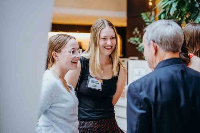 Two young women smiling and talking with an older man at an indoor event. One woman wears glasses and a light sweater, the other wears a black top and nametag. A background of plants and warm lighting is visible.