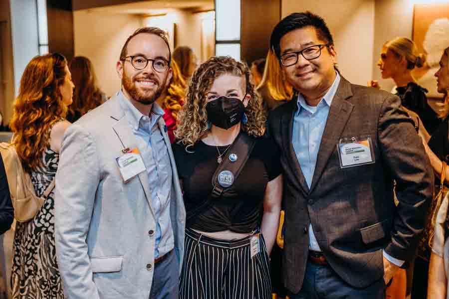 Three people, two men in suits and one woman wearing a black mask and striped pants, pose together and smile at an indoor event with other attendees socializing in the background.
