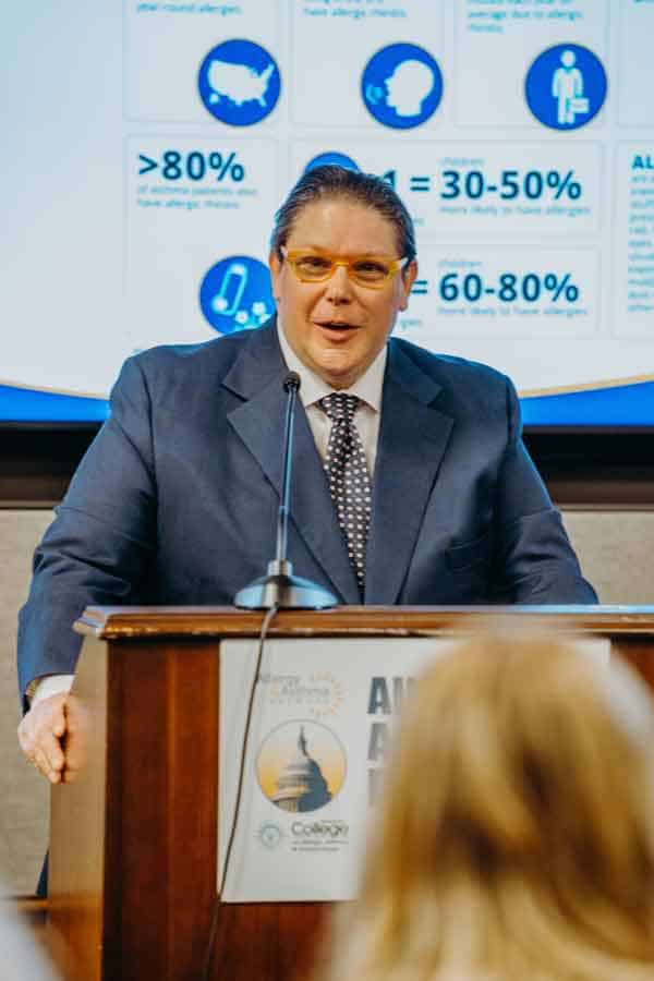 A man in a suit and tie speaks at a podium with a microphone. Behind him is a presentation slide displaying various infographics. An audience member is visible in the foreground.