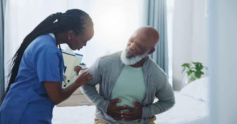 A nurse holding a clipboard speaks with an older man sitting on a bed and clutching his abdomen in pain, while the room is softly lit with natural light.