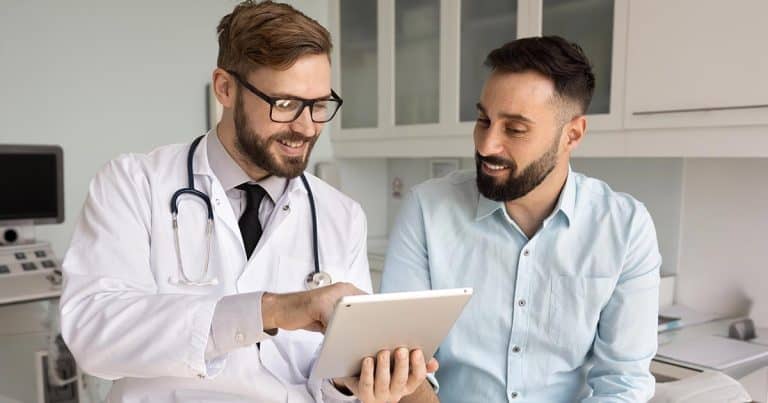 A doctor in a white coat and stethoscope shows information on a tablet to a smiling male patient in a light blue shirt, both sitting in a bright medical office.