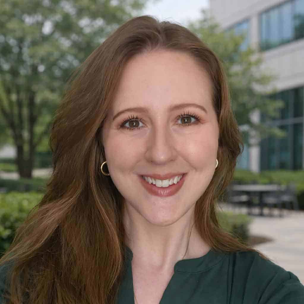 A woman with long, light brown hair smiles outdoors in front of a modern building with large windows and greenery, wearing a dark green top and hoop earrings.