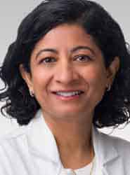 A woman with short, curly black hair is smiling and wearing a white coat against a light gray background.