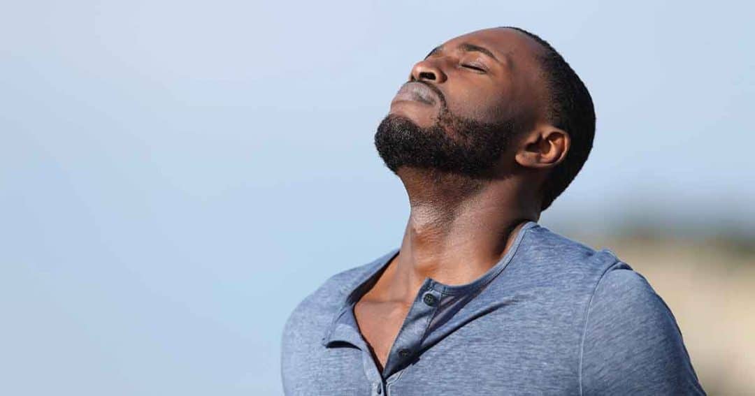 A man with a beard closes his eyes and tilts his head back, taking a deep breath outdoors. He wears a blue shirt and appears relaxed against a light, blurry background.