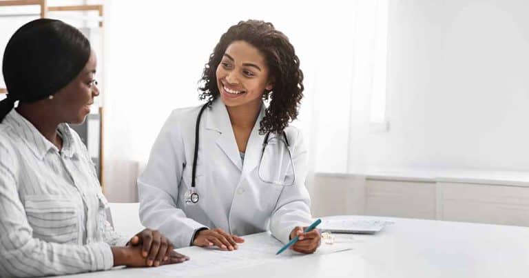 A female doctor in a white coat smiles while discussing paperwork with a female patient in an office setting. The doctor holds a pen and both appear engaged in conversation.