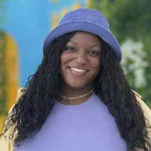 A smiling woman with long curly hair wears a purple bucket hat and a matching lavender top, standing outdoors with a blurred colorful background.