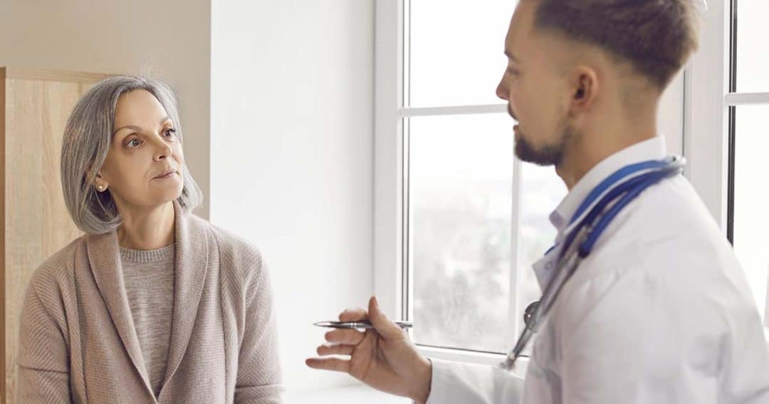 A middle-aged woman listens attentively to a male doctor with a stethoscope around his neck as he explains something, indoors near a window.
