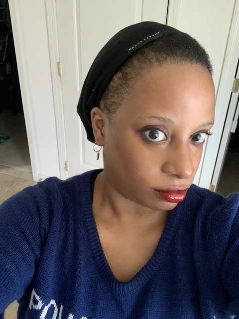 A person with short hair wearing a black headband, navy blue sweater, and red lipstick looks into the camera while taking a selfie indoors. White doors and walls are visible in the background.