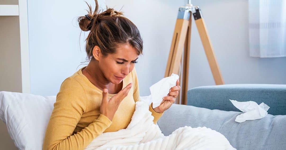 A woman sits on a couch covered with a blanket, holding a tissue and coughing or sneezing, appearing to be unwell in a home setting. Crumpled tissues are next to her.