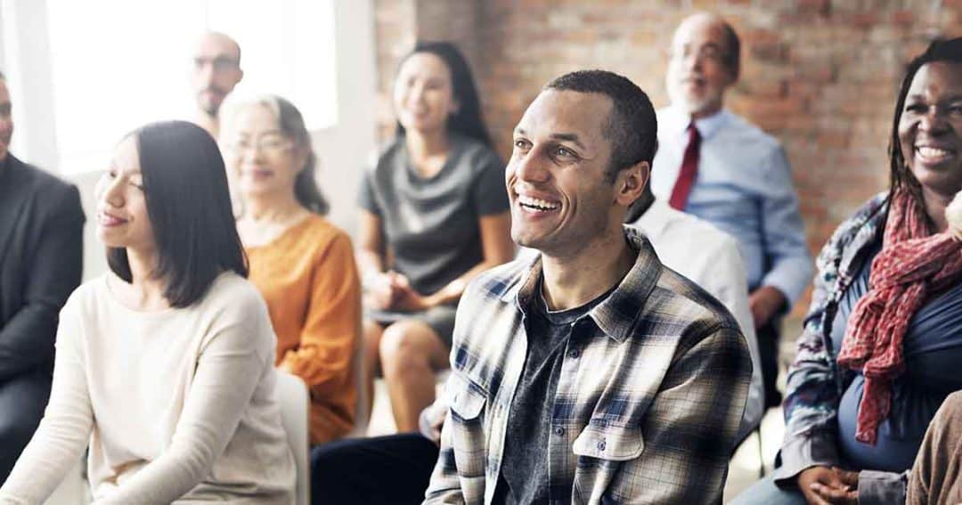 A diverse group of people sits closely together in a bright room, smiling and looking attentively toward the front, suggesting they are listening to a speaker or participating in an event.