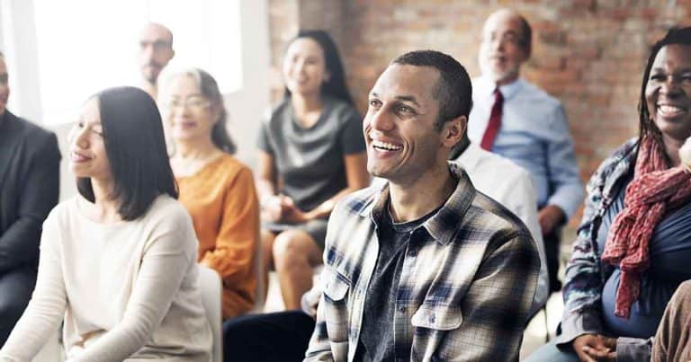 A diverse group of people sits closely together in a bright room, smiling and looking attentively toward the front, suggesting they are listening to a speaker or participating in an event.