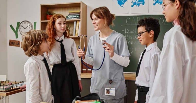 A nurse holding a stethoscope talks to four students in a classroom. The students wear school uniforms, and a chalkboard and bookshelf are visible in the background.