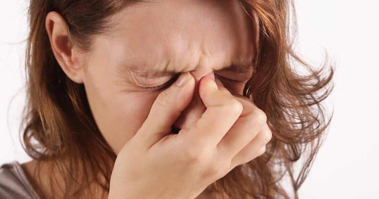 A woman with brown hair closes her eyes and pinches the bridge of her nose, appearing to be in discomfort or pain, possibly from a headache or sinus pressure, against a plain white background.