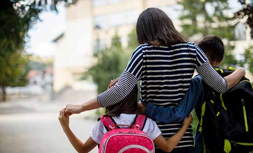 A woman with two children, all seen from behind, stands outdoors. She wears a striped shirt; the children have backpacks and hug her as they walk together.