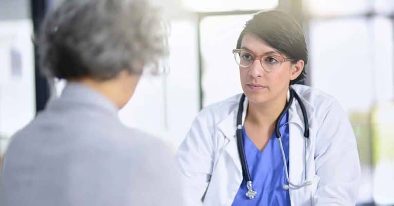 A doctor wearing a white coat and stethoscope attentively listens to an older patient during a medical consultation in a bright office.