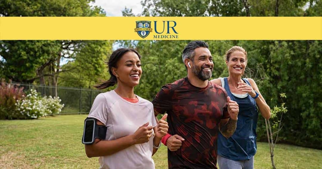 Three adults jog and smile together outdoors in a grassy park on a sunny day. Two women and one man appear active and happy. The UR Medicine logo is displayed at the top of the image.