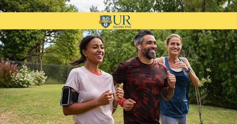 Three adults jog and smile together outdoors in a grassy park on a sunny day. Two women and one man appear active and happy. The UR Medicine logo is displayed at the top of the image.