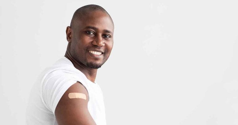 Smiling man in a white t-shirt with a bandage on his upper arm, standing against a plain white background.