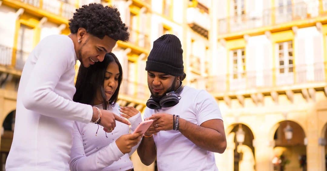Three young adults stand close together outside, smiling and looking at a smartphone held by the woman in the center. The background shows a sunny building with balconies.