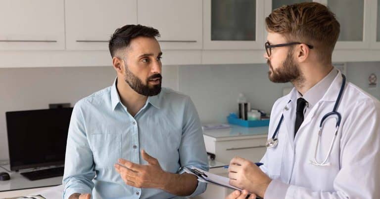A man in a light blue shirt talks to a doctor holding a clipboard in a medical office. The doctor listens attentively, wearing a white coat and stethoscope. Both appear engaged in their conversation.