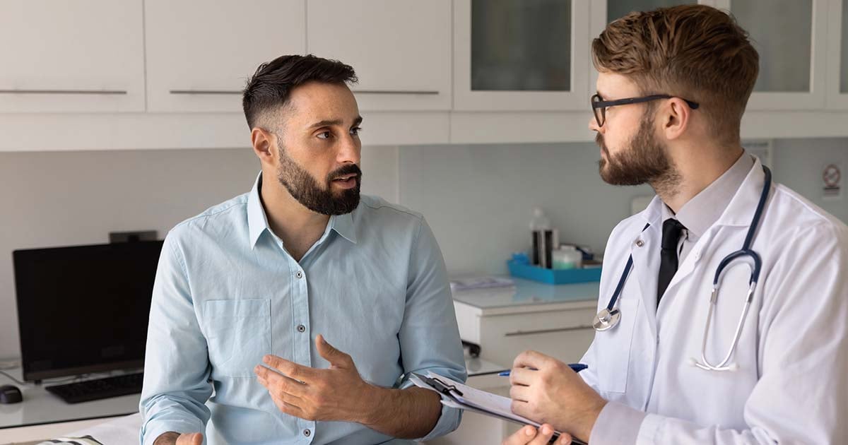 A man in a light blue shirt talks to a doctor holding a clipboard in a medical office. The doctor listens attentively, wearing a white coat and stethoscope. Both appear engaged in their conversation.