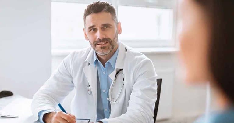 Male doctor in a white coat with a stethoscope sits at a desk, holding a pen and paper, attentively talking to a patient whose face is out of focus in the foreground. Bright office background.