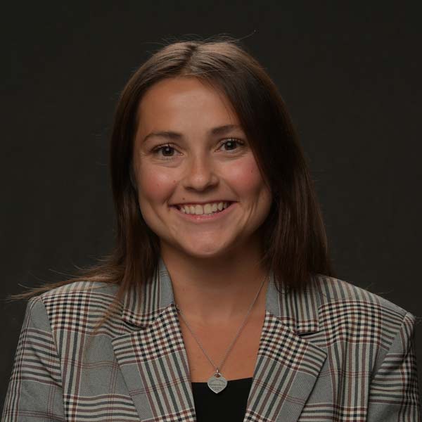 A young woman with brown hair smiles while wearing a plaid blazer and a heart-shaped pendant necklace.